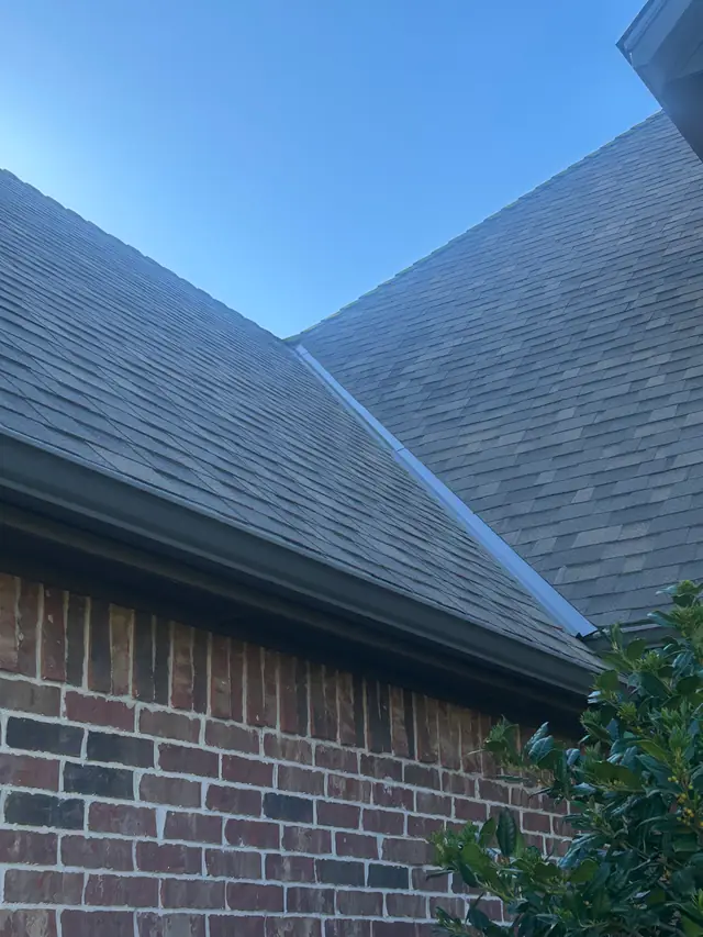 Gray shingle roof valley with metal flashing and a dark gutter along the eave above a brick wall