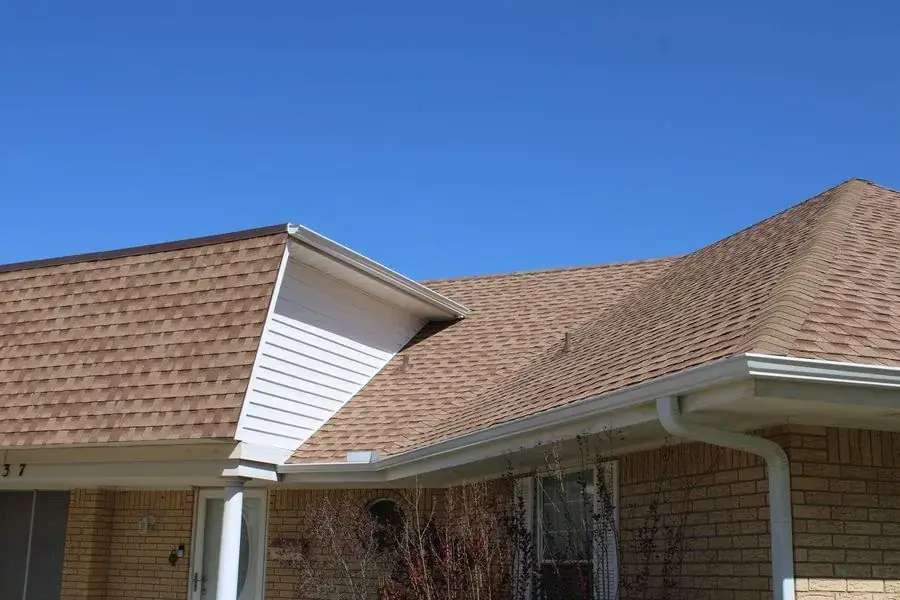 Tan shingle roof meeting a white-sided dormer with a white seamless gutter and downspout