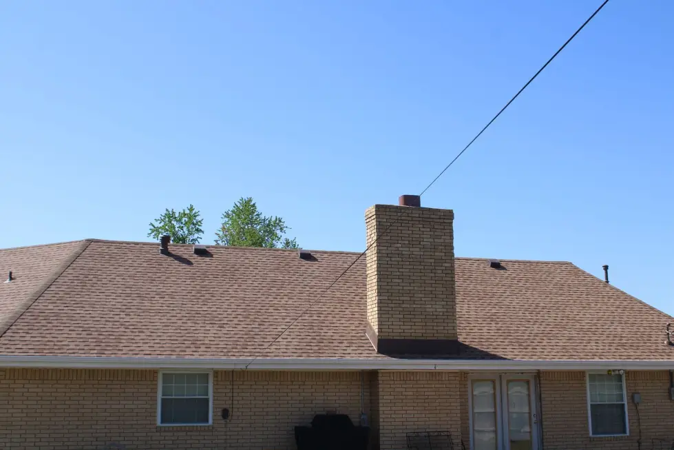 Brown shingle hip roof with a tan brick chimney over a brick ranch home