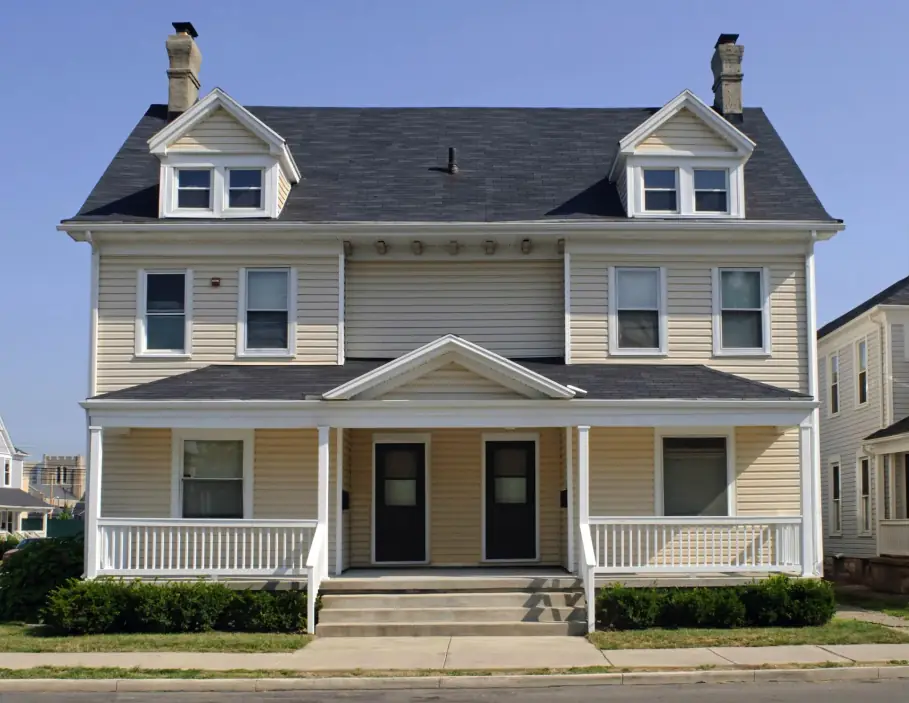 Two-story cream-sided house with a dark gray shingle roof, two gabled dormers and two brick chimneys